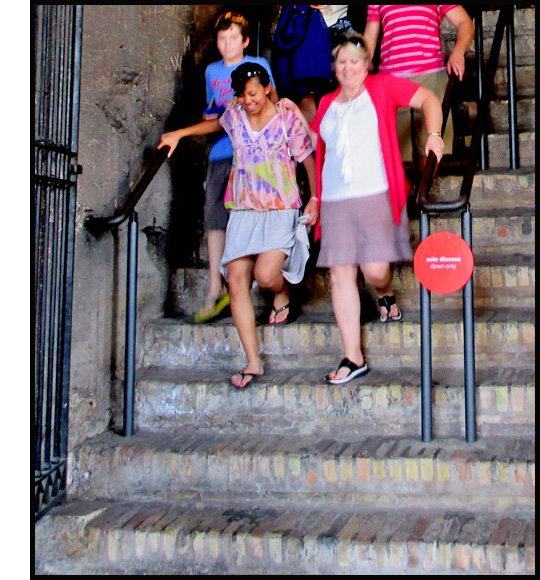 Photo shows people descending the left half of a stone or marble staircase.  Two girls in front of the crowd are giggling and each holding a railing as they step down.  They have reached the 4th stair from the bottom and are almost at the end of the railing.